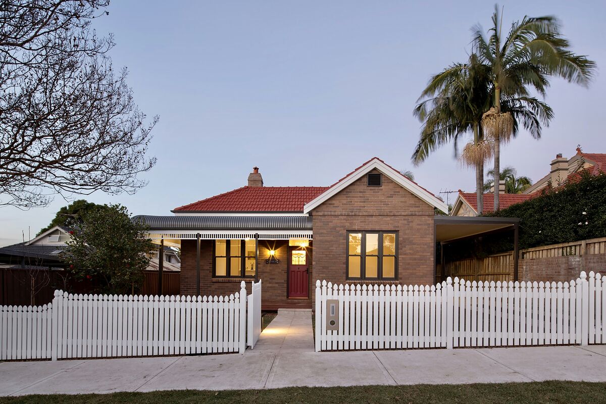 A renovated brick Federation home in Chatswood with a terracotta tiled roof, bullnose verandah and white picket fence, lights on at dusk