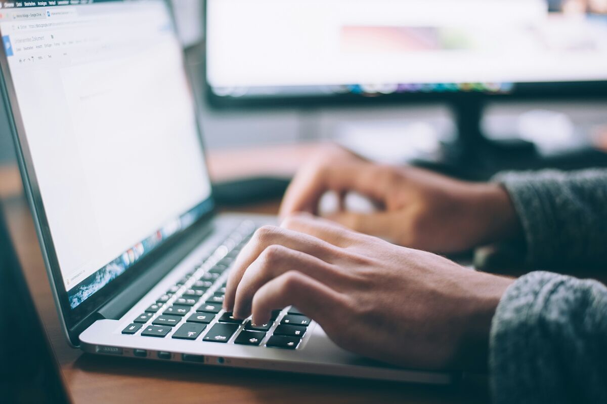 Close-up of hands typing on a laptop at a wooden desk with a second monitor softly out of focus behind