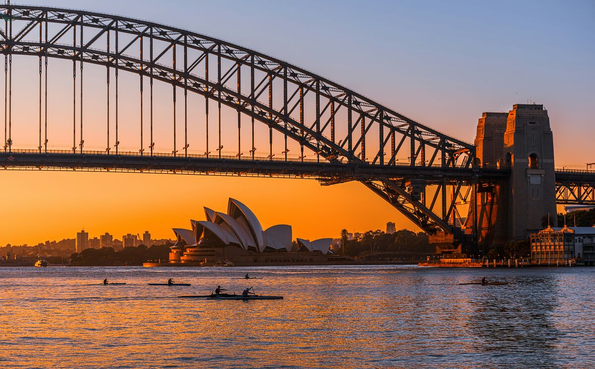 The Sydney Opera House framed under the arch of the Harbour Bridge at sunrise, with rowers on the harbour
