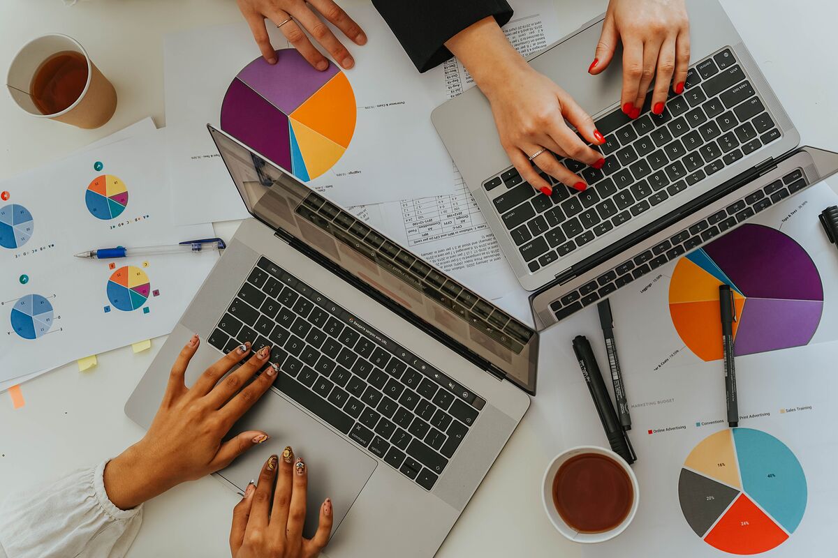 Overhead view of two people working on laptops at a white desk covered in printed marketing-budget pie charts and coffee cups